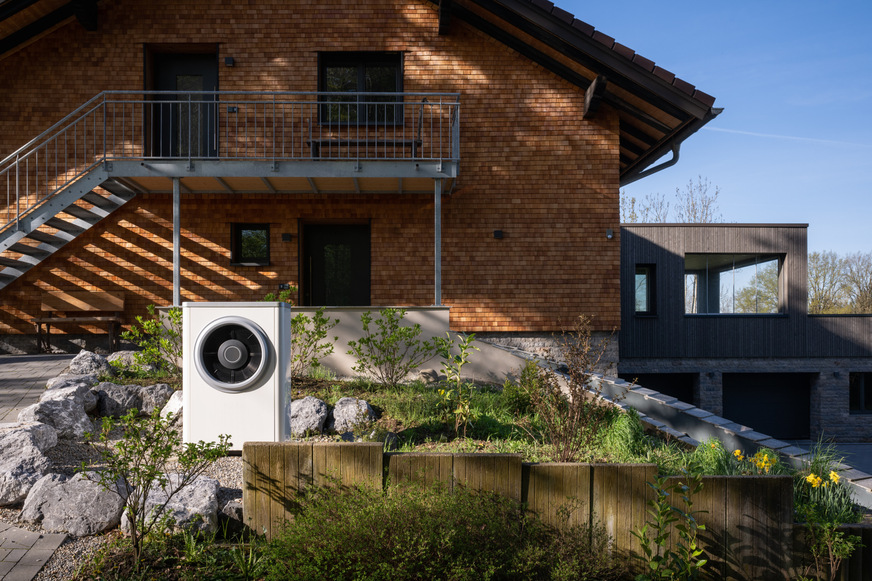 Wärmepumpe im Vorgarten eines Holzschindel-Hauses.