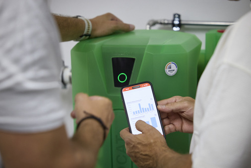 Zwei Personen prüfen Daten auf einem Smartphone vor einer grünen Maschine mit kreisförmiger Anzeige. - © Watercryst