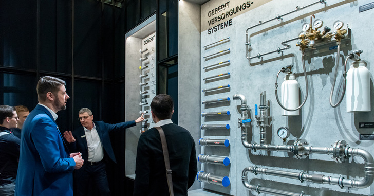 Alt-Text: „Gruppe von Fachleuten diskutiert die Integration digitaler Gebäudetechniklösungen und intelligenter Energiemanagement-Systeme anhand eines Plumbing-Displays mit Rohren und Fittings. - © Messe Frankfurt Alt-Text: „Gruppe von Fachleuten diskutiert die Integration digitaler Gebäudetechniklösungen und intelligenter Energiemanagement-Systeme anhand eines Plumbing-Displays mit Rohren und Fittings. - © Messe Frankfurt