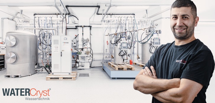 Ein lächelnder Mann steht vor einer industriellen Wasseraufbereitungsanlage in einer hellen, sauberen Einrichtung. - © Watercryst