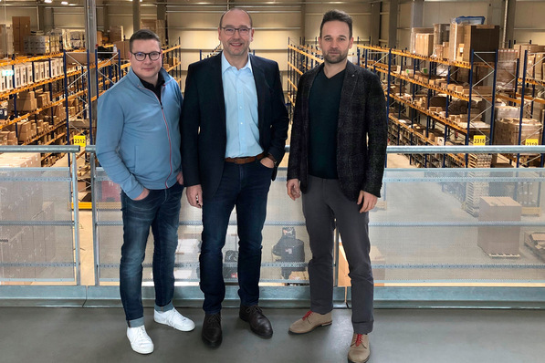 Alt-Text: „Drei Männer diskutieren in einem Lagerraum voller digitaler Gebäudetechnik-Komponenten und Smart-Building-Lösungen, umgeben von Regalen mit elektronischen Geräten und Bauteilen. - © Empur