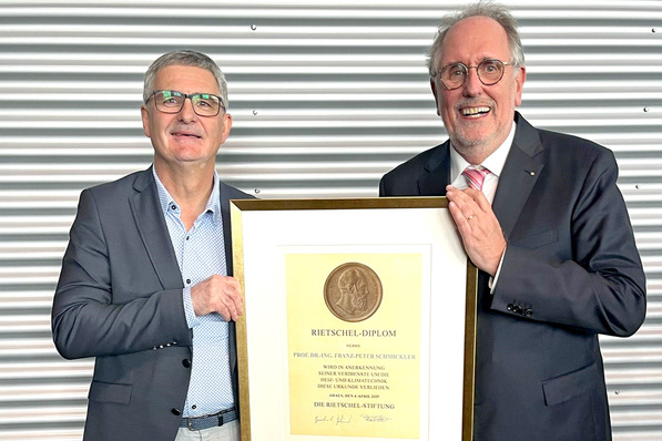 „Zwei lächelnde Männer präsentieren stolz ein eingerahmtes Zertifikat, das für Innovation und Exzellenz in der Sanitär-, Heizungs- und Klimatechnik ausgezeichnet wurde, vor einer Wand aus welligem Metall. - © Stefan Tuschy / BTGA