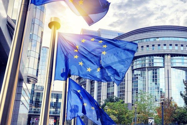 Alt-Text-Vorschlag: „Mehrere EU-Flaggen vor einem modernen Bürogebäude, Symbolbild für europäische Normen und Richtlinien in der technischen Dämmung und Gebäudetechnik.“. - © Symbiot - stock.adobe.com